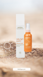 Aveda Daily Light Guard SPF 30 sunscreen and Aveda Sun Care Protective Hair Veil bottles are displayed on a blurred sandy background. - Ihloff Salon and Day Spa