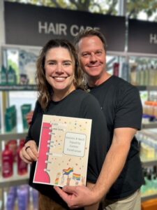 Two people holding a fundraising poster for Dennis R. Neill Equality Center, with a goal thermometer and colorful decor, standing in front of hair care products. Both are smiling at the camera.