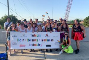 A group photo of people outdoors holding small pride flags and a sign that reads "Tulsa's Hair, Beauty, Wellness industry" with various business names and logos. Construction cranes and sky in the background.