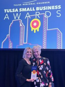 Two people are standing together holding an award in front of a "Tulsa Small Business Awards" sign at an event. The background shows a graphic skyline and the text "TULSA REGIONAL CHAMBER.