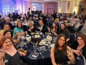 A group of people sitting at a round table at a formal event, raising their glasses in a toast. The room is filled with other guests seated at tables.