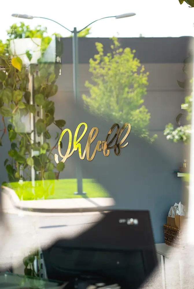 Gold lettering spelling "Ohloff" is seen on a glass window, hinting at a salon inside, with outdoor greenery and a street reflected in the glass. - Ihloff Salon and Day Spa