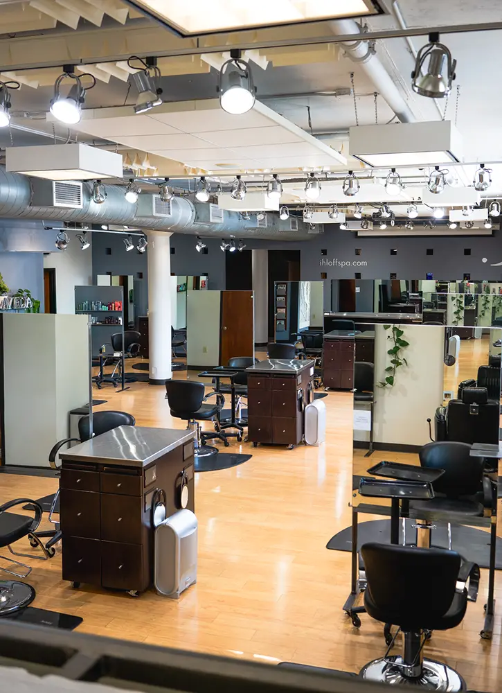 A modern hair salon and spa interior featuring empty styling stations, black chairs, mirrors, wooden floors, and overhead lighting—perfect for your next hair style or facial. - Ihloff Salon and Day Spa