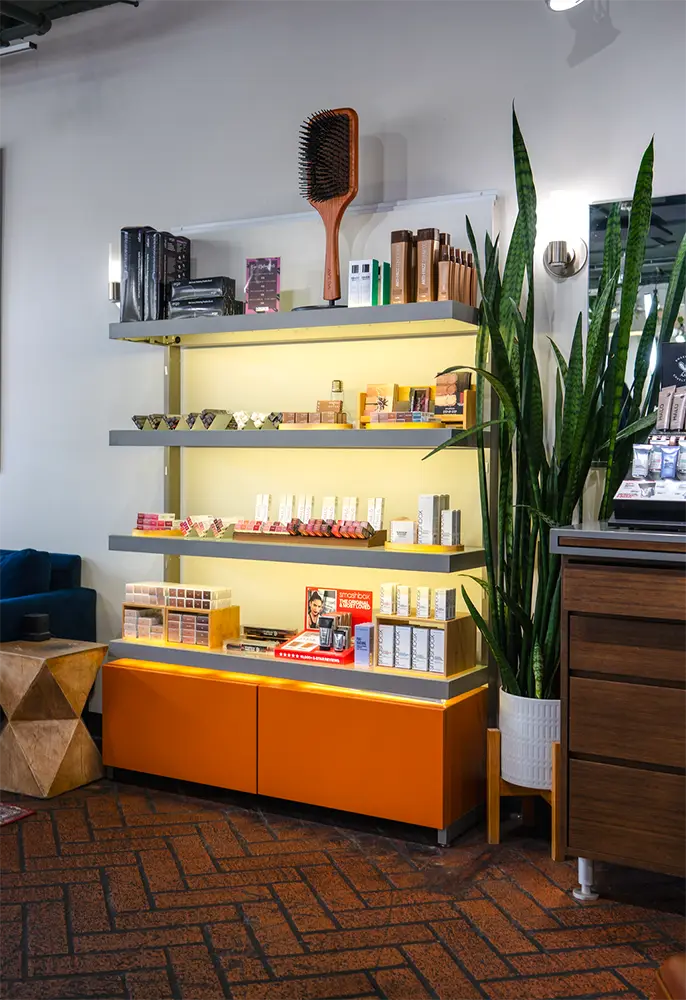 A display shelf with various beauty products, facial essentials, a large hairbrush, a tall potted plant, and an orange cabinet in a modern salon or retail space. - Ihloff Salon and Day Spa