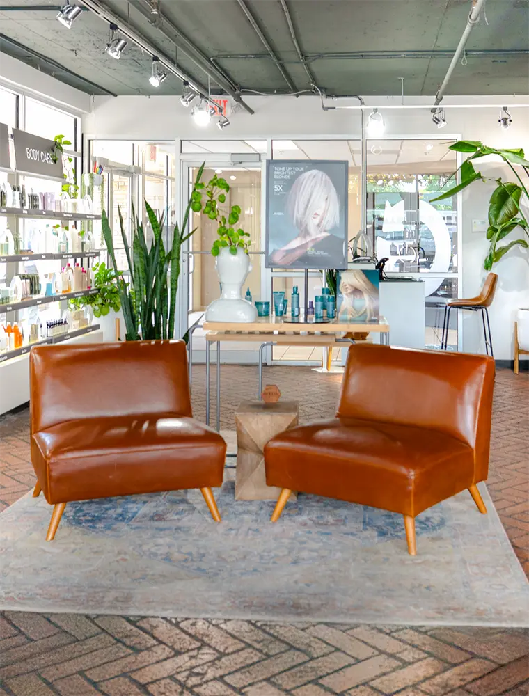 Two brown leather chairs face each other on a rug in a modern salon lobby, with lush plants, shelves of hair style products, and a table in the background. - Ihloff Salon and Day Spa