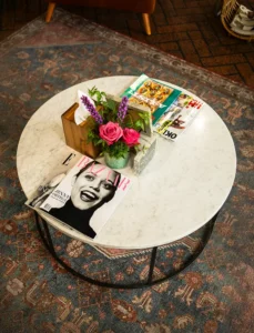 A round marble coffee table with magazines, a small vase of pink and purple flowers, and a wooden box sits on a patterned rug, creating a spa-like ambiance perfect for a salon waiting area or hair color consultation space. - Ihloff Salon and Day Spa