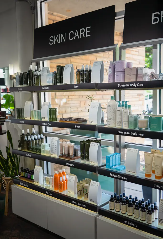 Shelves display various skin care and body care products, organized by category, in a well-lit salon with large windows, plants nearby, and hints of hair style inspiration throughout the space. - Ihloff Salon and Day Spa