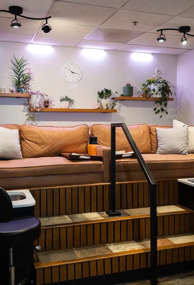 A cozy salon seating area with tan sofas, white and beige pillows, wooden steps, potted plants on wall shelves, a white clock, and soft overhead lighting—perfect for relaxing before your hair color or facial appointment. - Ihloff Salon and Day Spa