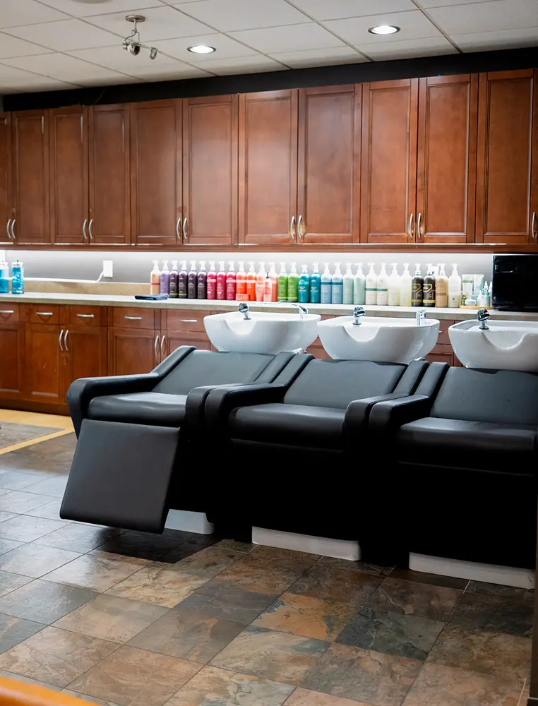 Three black salon shampoo chairs face white sinks, with bottles of hair products and vibrant hair color arranged on the countertop against wooden cabinets in a well-lit salon. - Ihloff Salon and Day Spa