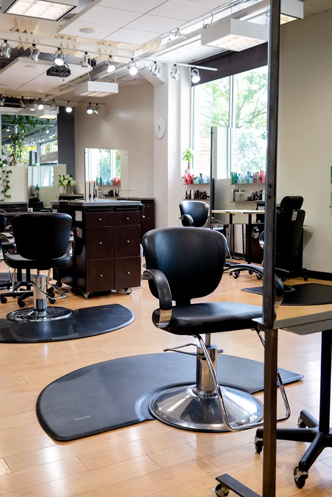 A modern hair salon interior with empty black styling chairs, mirrors, cabinets, and hair products on display, featuring wooden floors and large windows letting in natural light—perfect for your next hair style or vibrant hair color. - Ihloff Salon and Day Spa