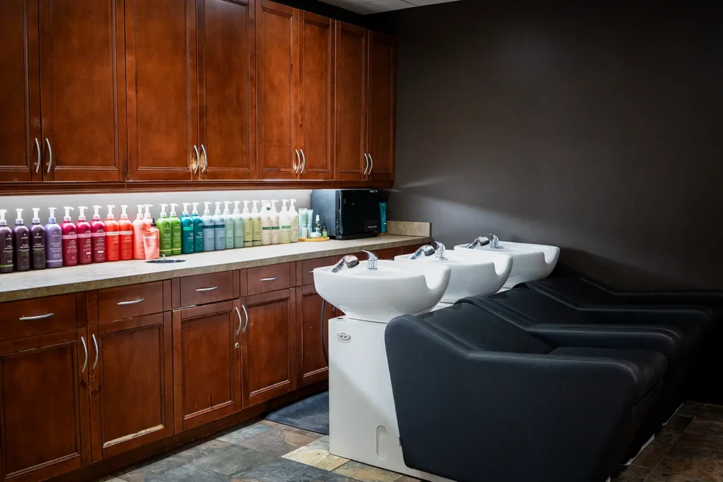 Three salon shampoo stations with black chairs are positioned in front of wooden cabinets, with rows of colorful hair product bottles on the counter—creating a relaxing spa atmosphere perfect for any hair style. - Ihloff Salon and Day Spa