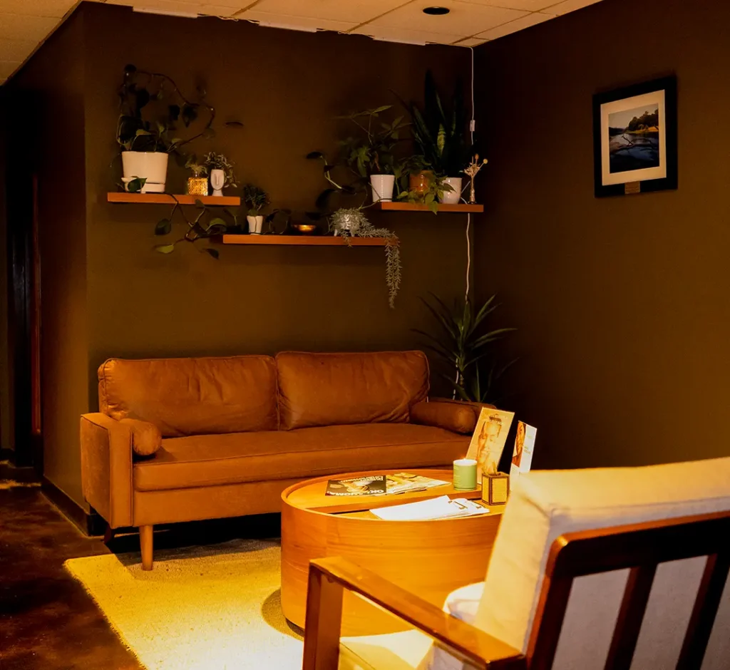 A cozy salon-inspired seating area with a brown sofa, a wooden coffee table, a cream chair, wall shelves with potted plants, and a framed picture on a dark brown wall. - Ihloff Salon and Day Spa