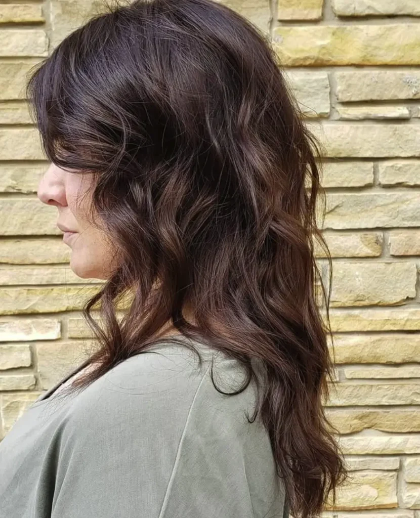 Woman with wavy, shoulder-length dark brown hair color standing in front of a tan brick wall, shown in profile. - Ihloff Salon and Day Spa