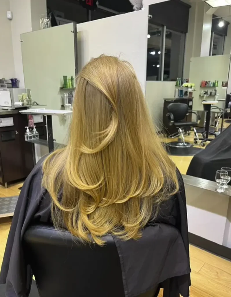 Person with long, layered blonde hair sits in a salon chair, facing away from the camera; salon mirrors, spa chairs, and hair products visible in the background. - Ihloff Salon and Day Spa