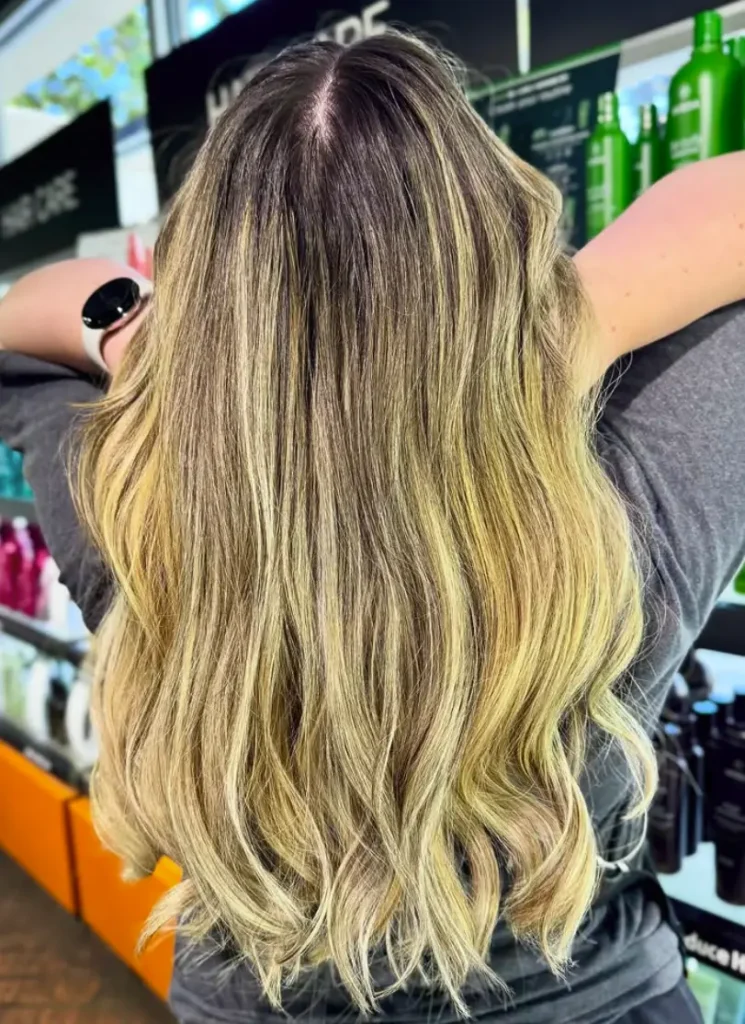 A person with long, wavy, blonde hair stands in front of shelves with hair care products at a salon, facing away from the camera. - Ihloff Salon and Day Spa