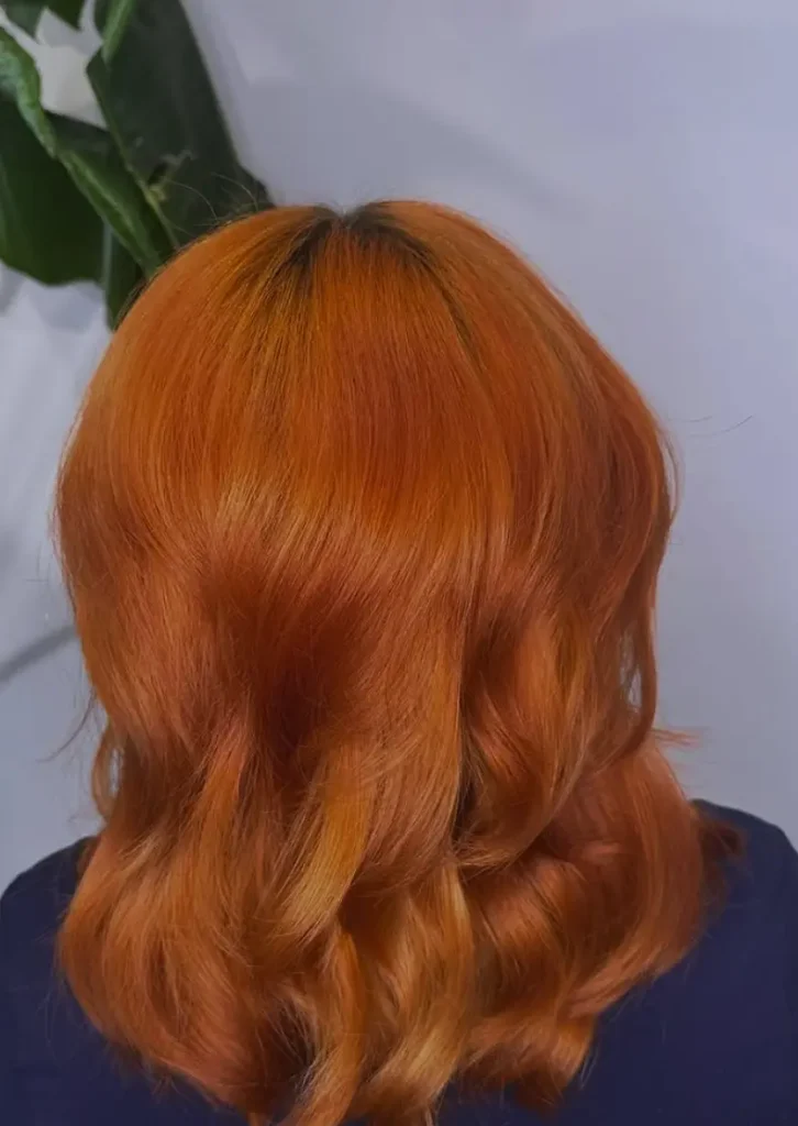 Person with a shoulder-length, wavy hair style in bright orange hair color shown from the back against a plain background, with a green plant partially visible. - Ihloff Salon and Day Spa